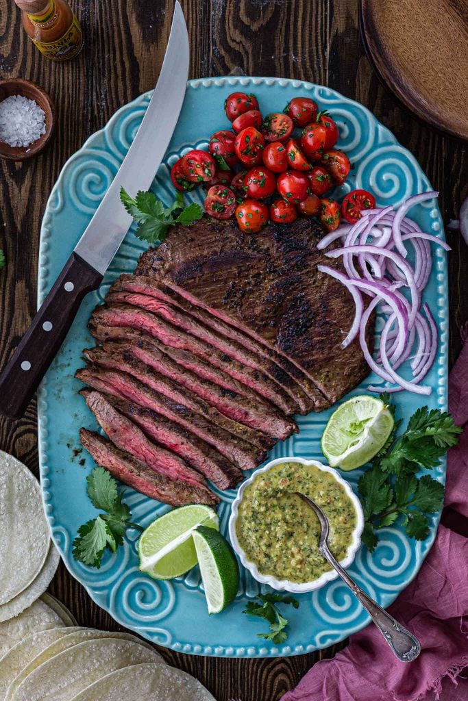 A platter of carne asada with tomato salsa, lemon wedges, cilantro, sliced onions, tortillas and sauce.