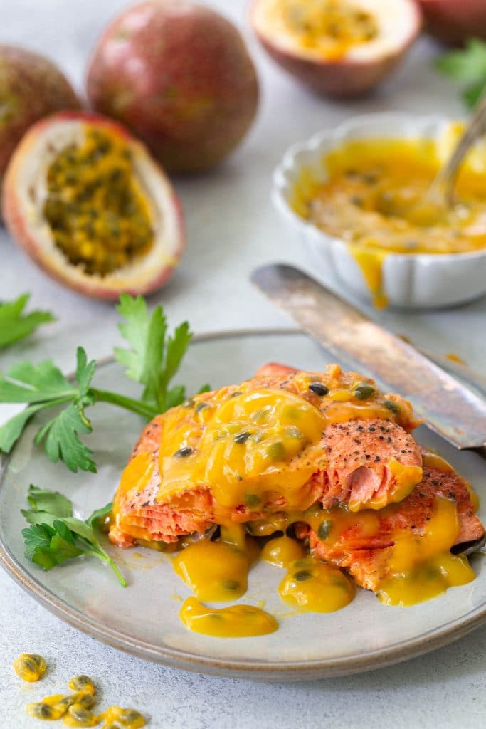 Two pieces of pan fried salmon with passion fruit sauce in a small plate, garnished with fresh parsley. The sauce and fresh passion fruits can be seen in the back.