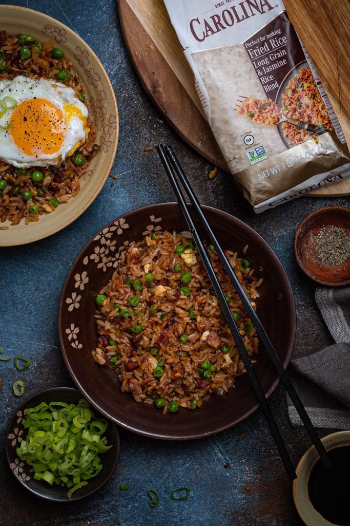 Two bowls of bacon fried rice, a smaller bowl with green onions, one with soy sauce and one with black pepper. You can also see a bag of Carolina rice on a cutting board.