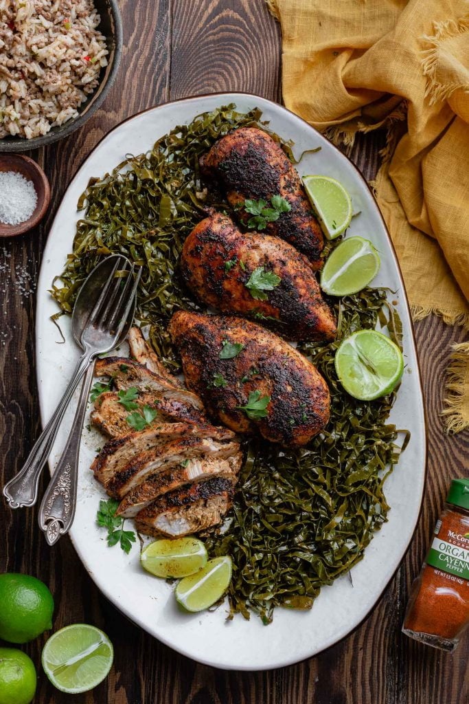 A platter of blackened chicken breasts over a bed of collard greens. You can see lime wedges and halves as a garnish, a bowl of dirty rice, a bowl of salt, a yellow napkin and a jar of cayenne pepper.