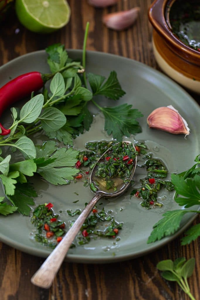 A plate with chimichurri, herbs, a garlic cloves and a chili pepper.