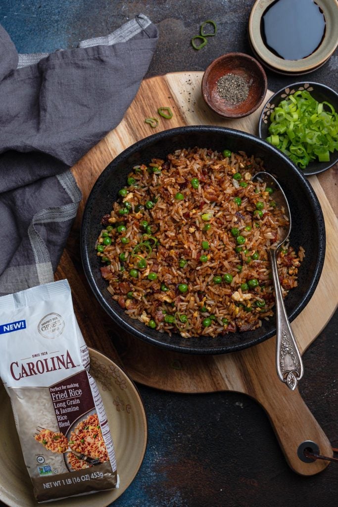 A serving bowl with bacon fried rice, 3 smaller bowls with toppings (scallions, soy sauce and black pepper), a napkin and a bag of Carolina fried rice blend.
