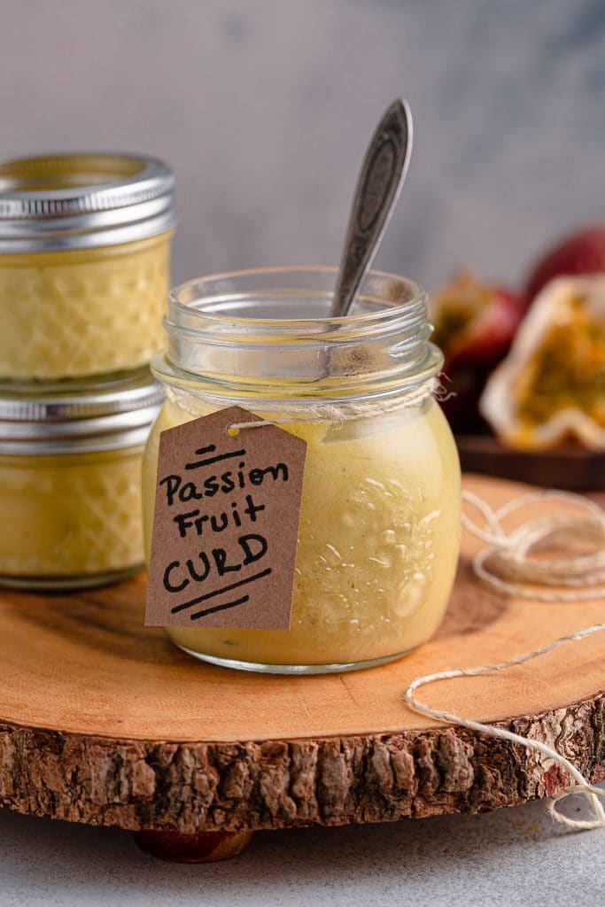 A jar of passion fruit curd with a spoon inside. Two more jars and some passion fruit can be seen in the background.