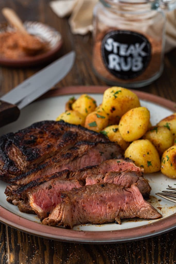 A perfectly cooked dry rubbed steak with baby potatoes. You can see a jar of steak rub in the background.