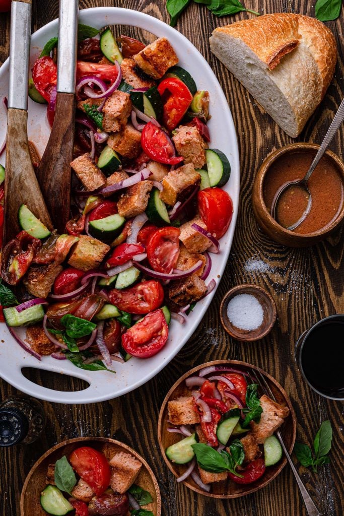 A platter of panzanella salad and two plates of individual servings. You can see the Italian vinaigrette in a bowl on the side and a smaller bowl with salt.