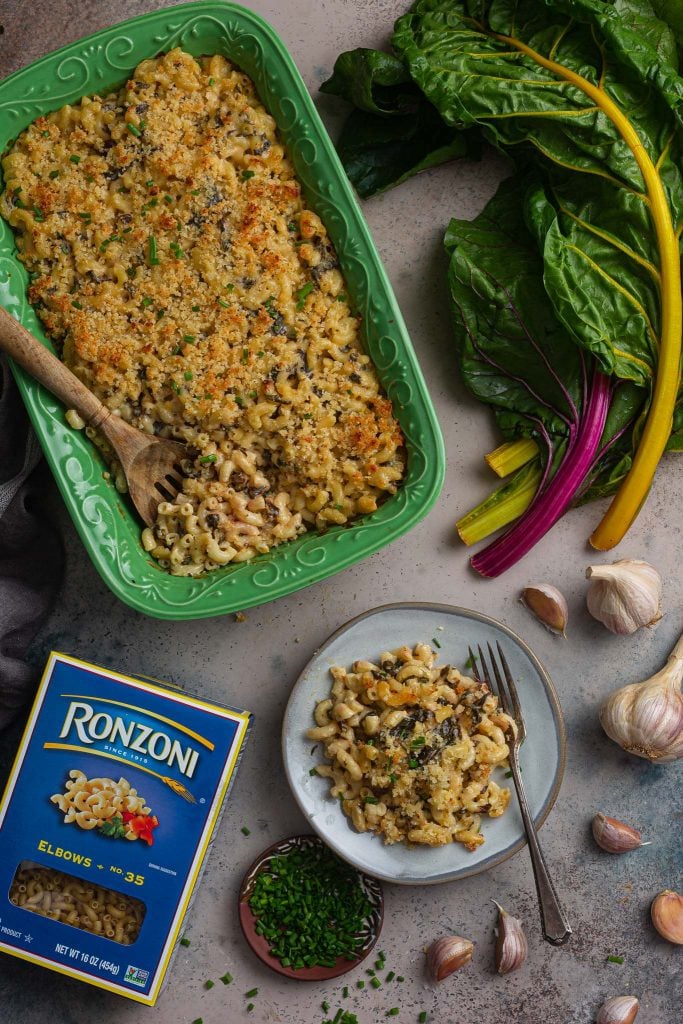 A serving scene. You can see the baking dish with roasted garlic mac and cheese, a plate with an individual portion, a Ronzoni box, a little plate with chives (for garnishing), some garlic and a bunch of Rainbow Chard.