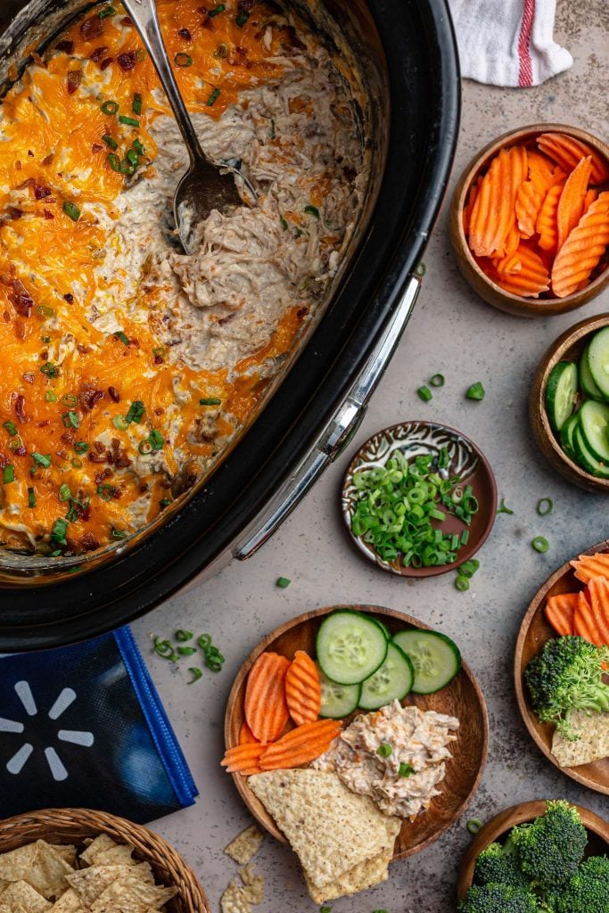 A serving scene: the slow cooker with the cheesy chicken dip, bowls with sliced carrots, cucumbers, broccoli and green onions, and 2 plates with individual servings.