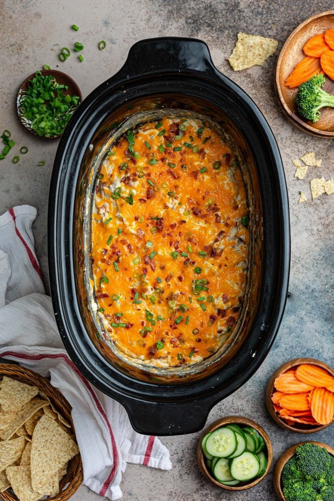 An overhead photo of a slow cooker with cheesy ranch chicken dip. Chips and vegetables on the side.