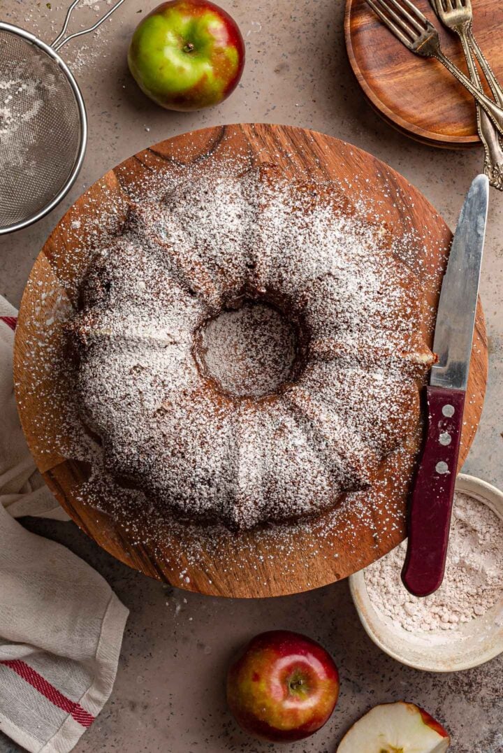 An overhead photo of the cake on a cake stand. You can also see a dish towel, a knife, a bowl with powdered sugar, a strainer, some apples and a few plates and utensils.