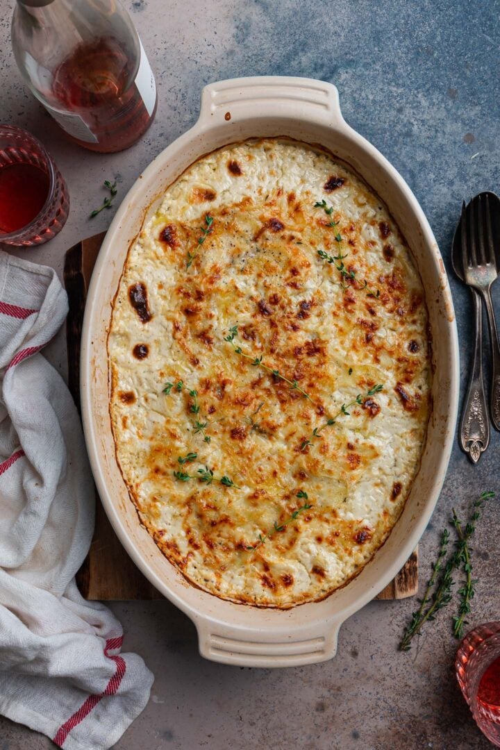 An overhead photo of Potatoes Dauphinoise dish on a wood board. You can also see a bottle of rose wine, two glasses, some thyme sprigs, utensils and a dish towel.