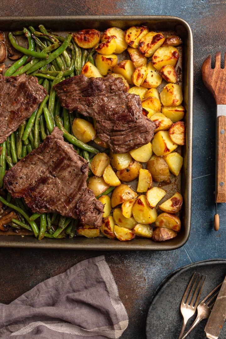 A serving scene: sheet pan steak, veggies, plates, forks and a serving utensil.