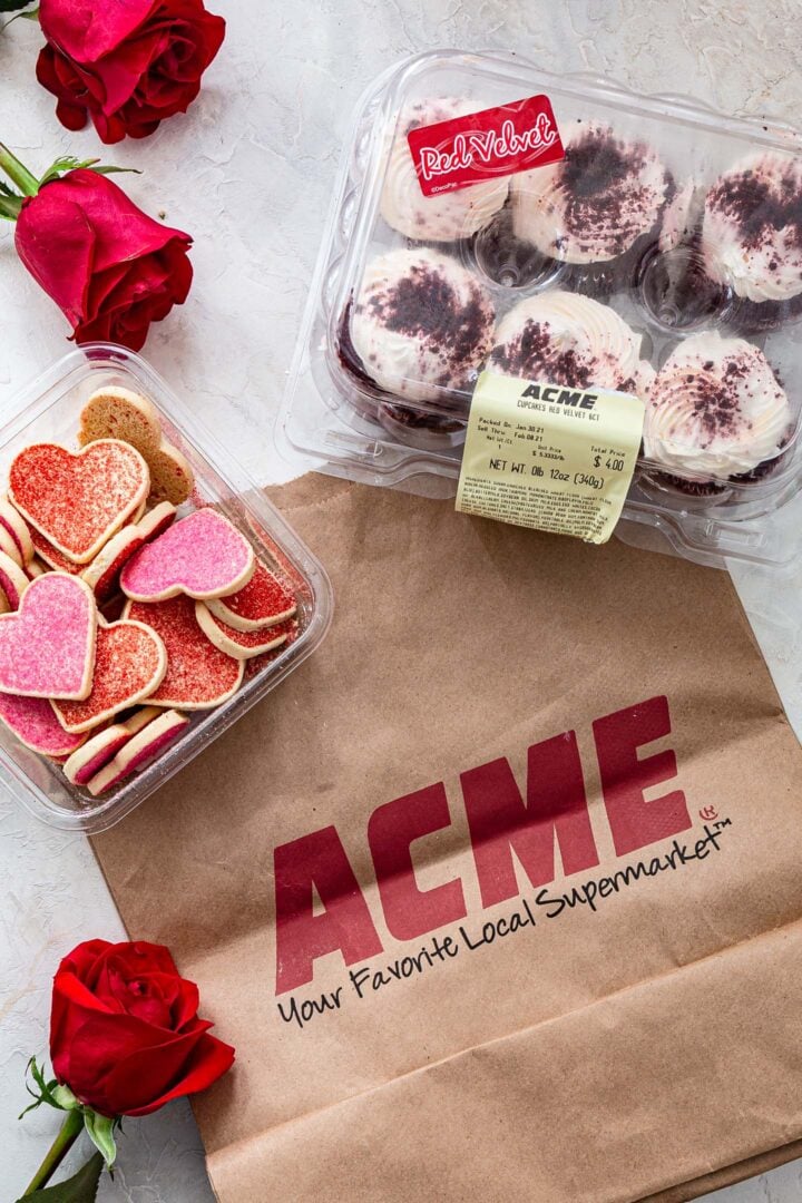 An ACME bag, a container of Red Velvet cupcakes, a container of heart shaped cookies and some roses.