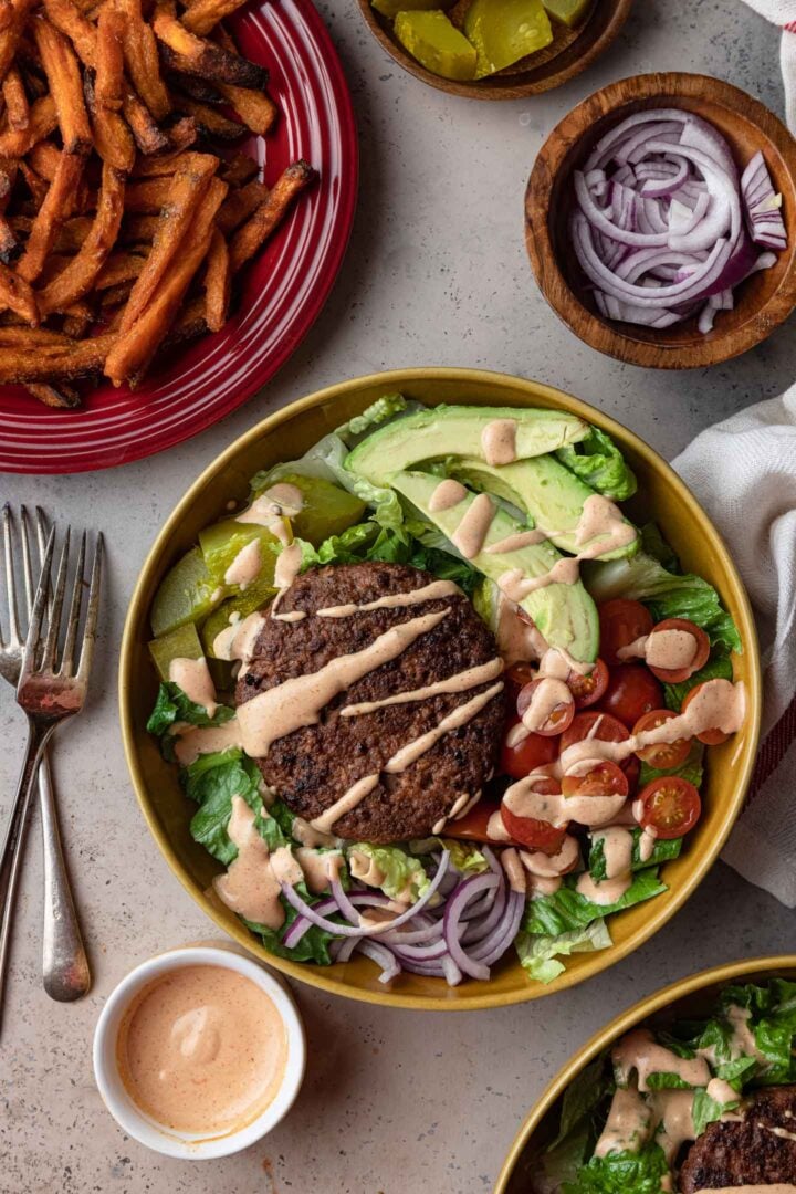 An overhead image of two veggie burger bowls, drizzled with special sauce and served with sweet potato fries and more sauce on the side.