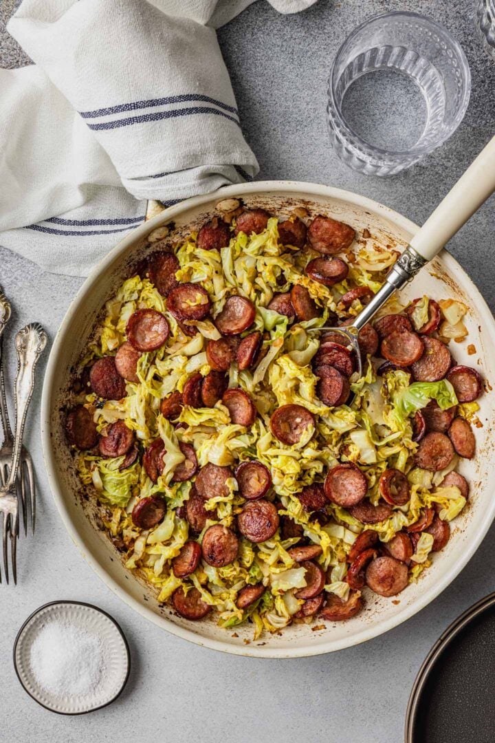 Sausage and Cabbage Skillet, a few forks, plates, a glass of water and a small pinch bowl with salt.