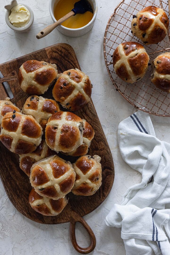 An overhead photo of some hot cross buns on a wire rack, some on a wood board and a kitchen towel.