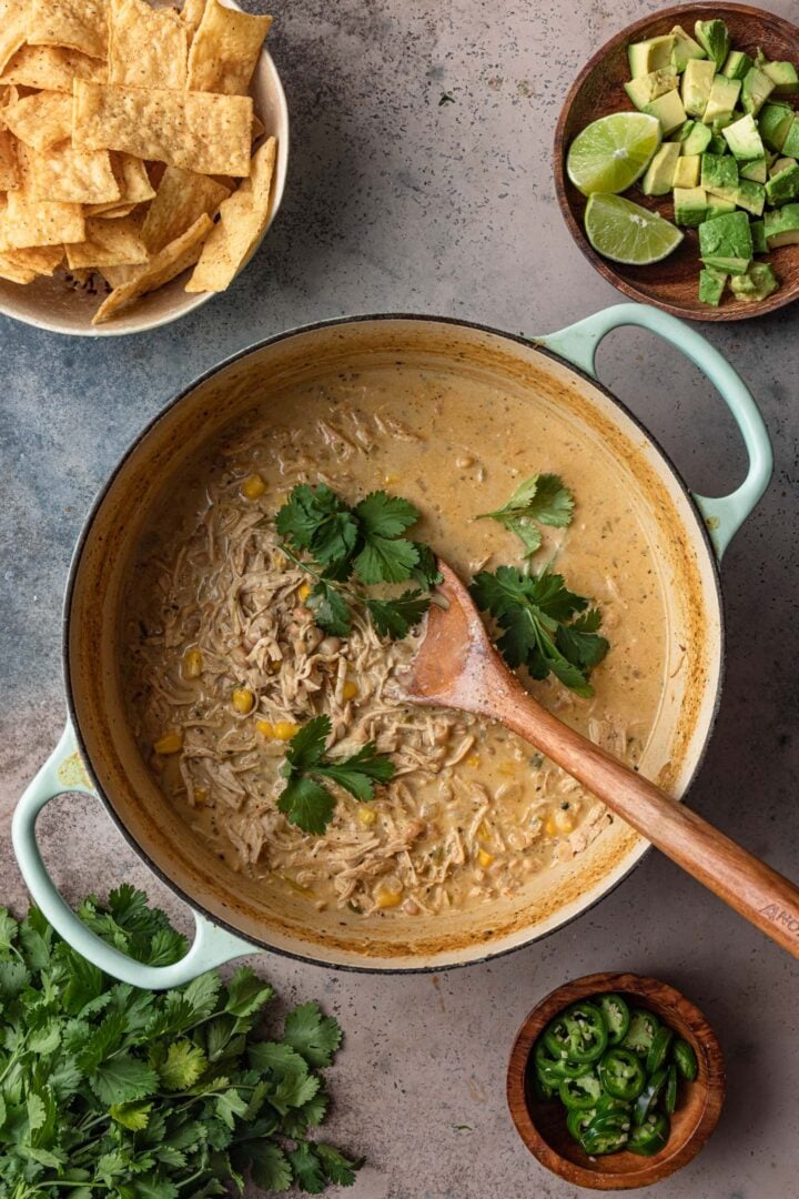 A photo of a Dutch Oven with white chicken chili. Toppings are on the side: jalapeños, cilantro, avocado and lime wedges, tortilla strips.