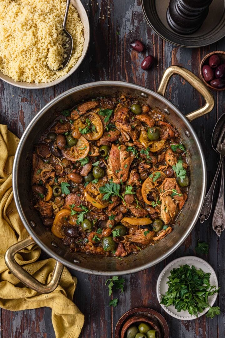 An overhead photo of a pan of chicken tagine served with a bowl of couscous. You can also see a kitchen towel, a small bowl with parsley leaves, bowls with olives and utensils.