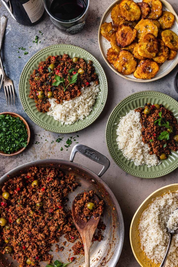 Serving scene. A pan of picadillo, along with two plates of picadillo served with rice. A bowl of white rice and a plate of tostones are also in the shot.