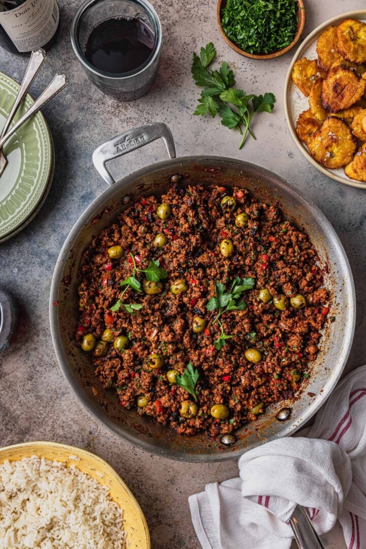 An overhead photo of a skillet of picadillo with rice and tostones served on the side along with red wine, parsley, plates, and a kitchen cloth.