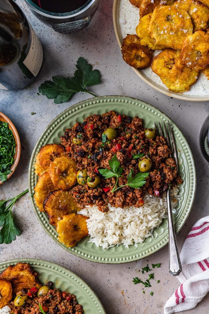 An individual serving of beef picadillo with white rice and tostones. A plate of tostones. a bottle of wine, two wine glasses, chopped parsley and a kitchen towel are on the side.