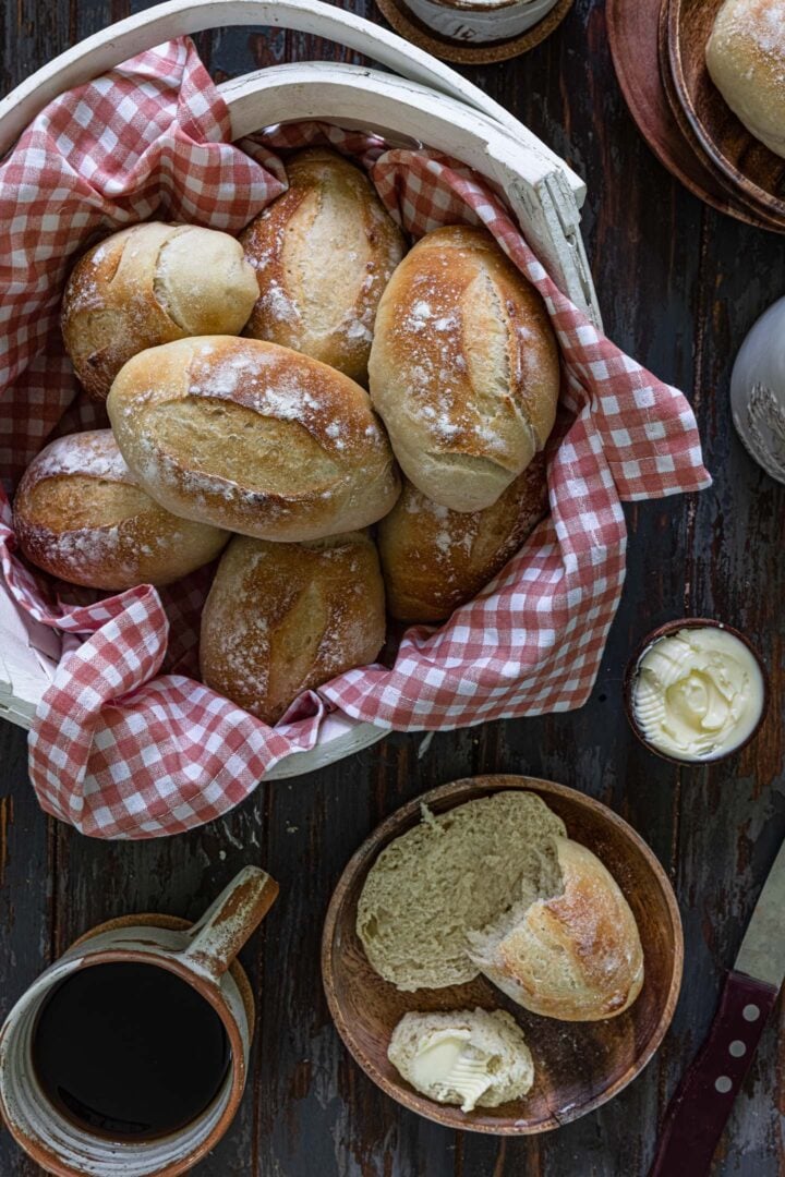 An overhead photo of a basket of pão francês, a coffee mug and one of the rolls split open to show the soft crumb.