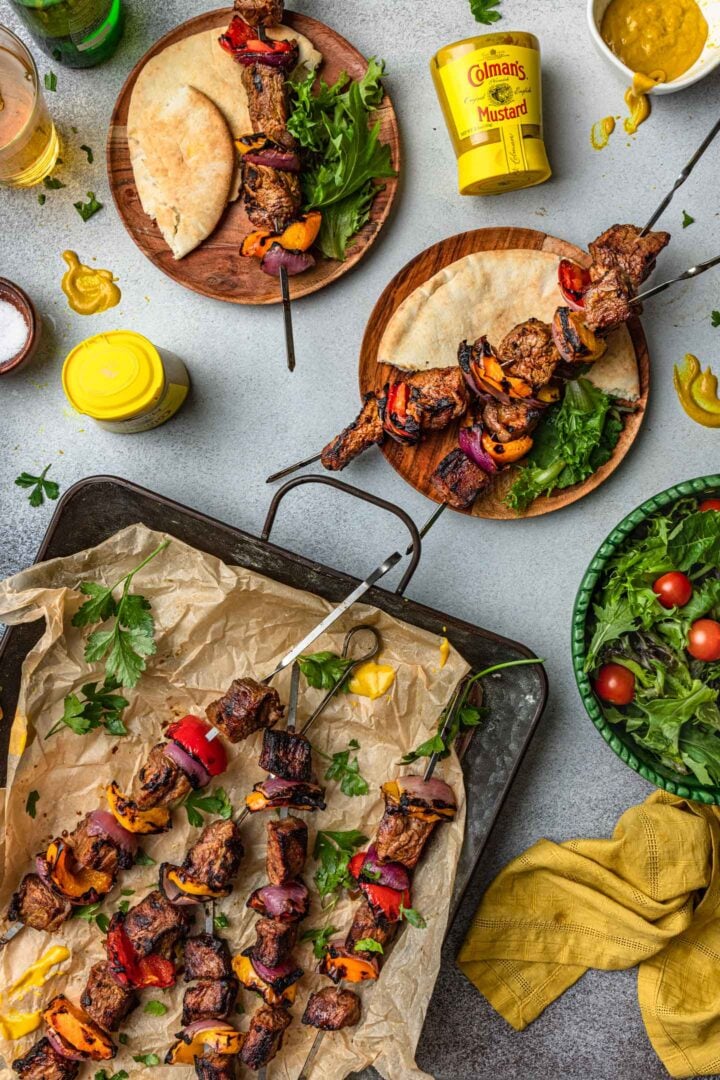 Marinated steak kabobs on a tray, and also some kabobs on individual plates, served with salad and pita bread.