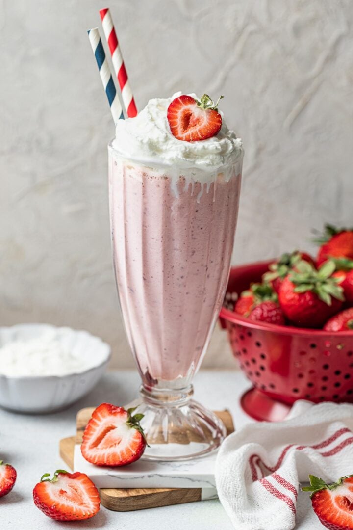 A glass of strawberry milkshake, topped with whipped cream and a strawberry, with two striped straws. 