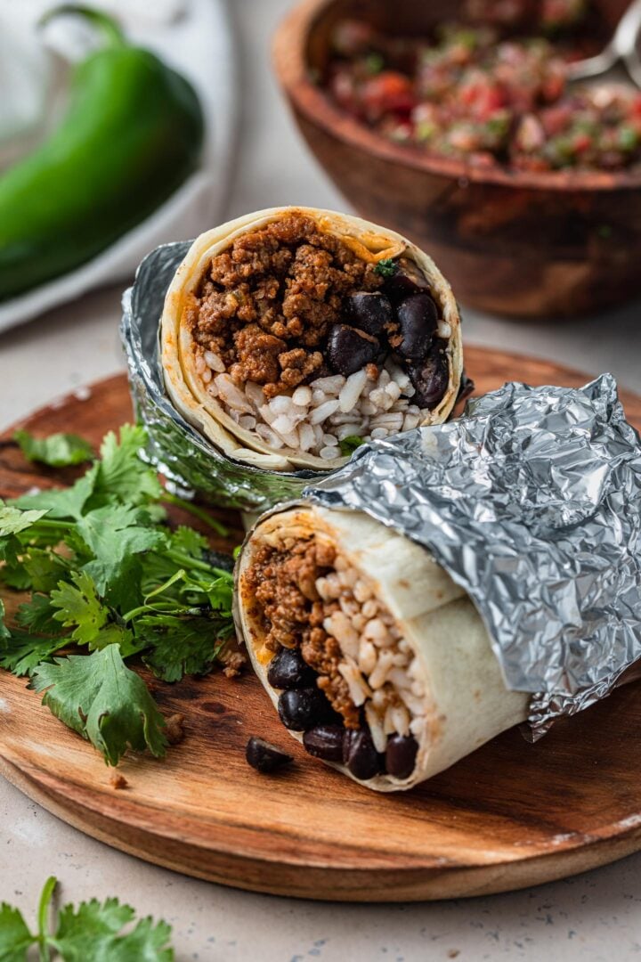 A cut ground beef burrito wrapped in foil, on a plate with cilantro.