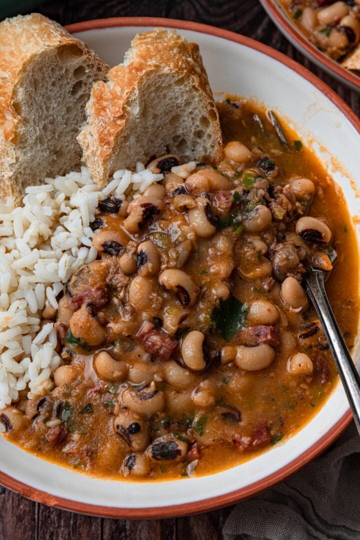 A close up of a serving of black-eyed peas in a bowl, with white rice and bread.