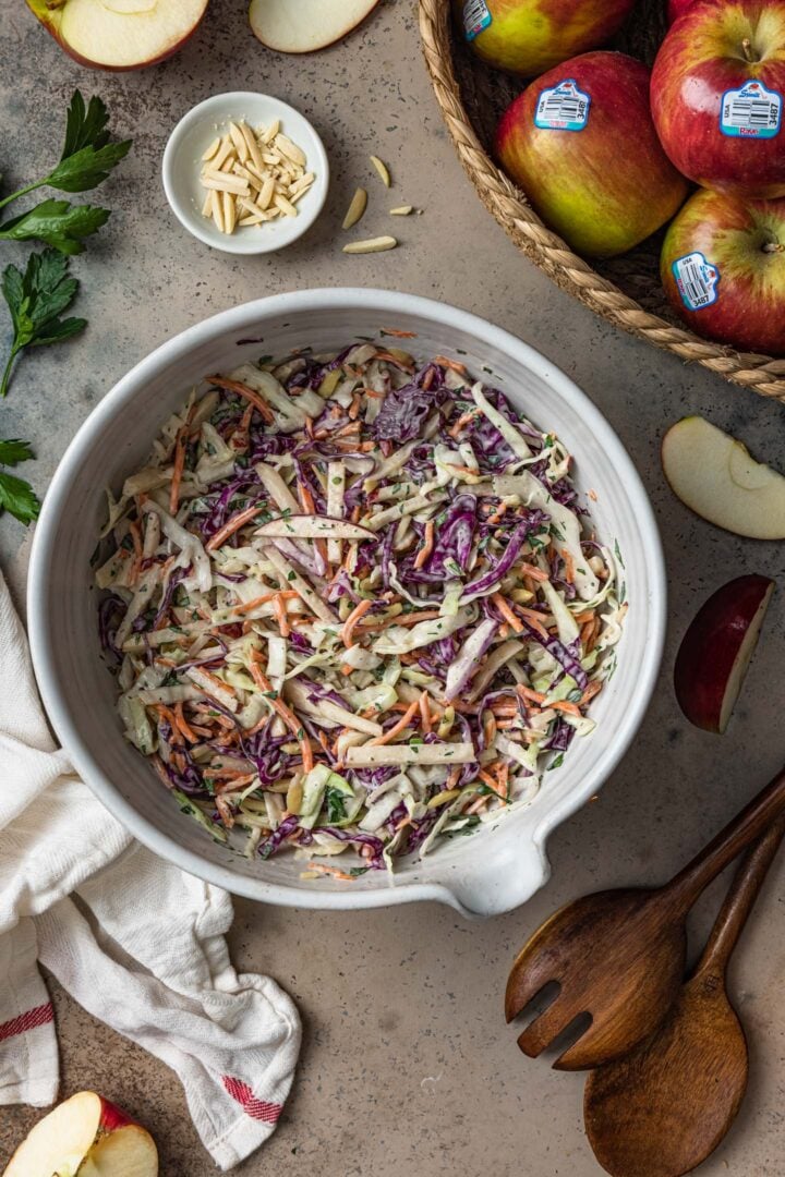 A bowl of apple slaw, salad servers, a kitchen towel, a small bowl of slivered almonds and a basket full of apples.