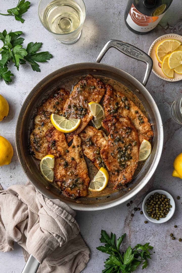 A pan with chicken piccata. On the side you can see some parsley, a few glasses of white wine, capers and lemons.