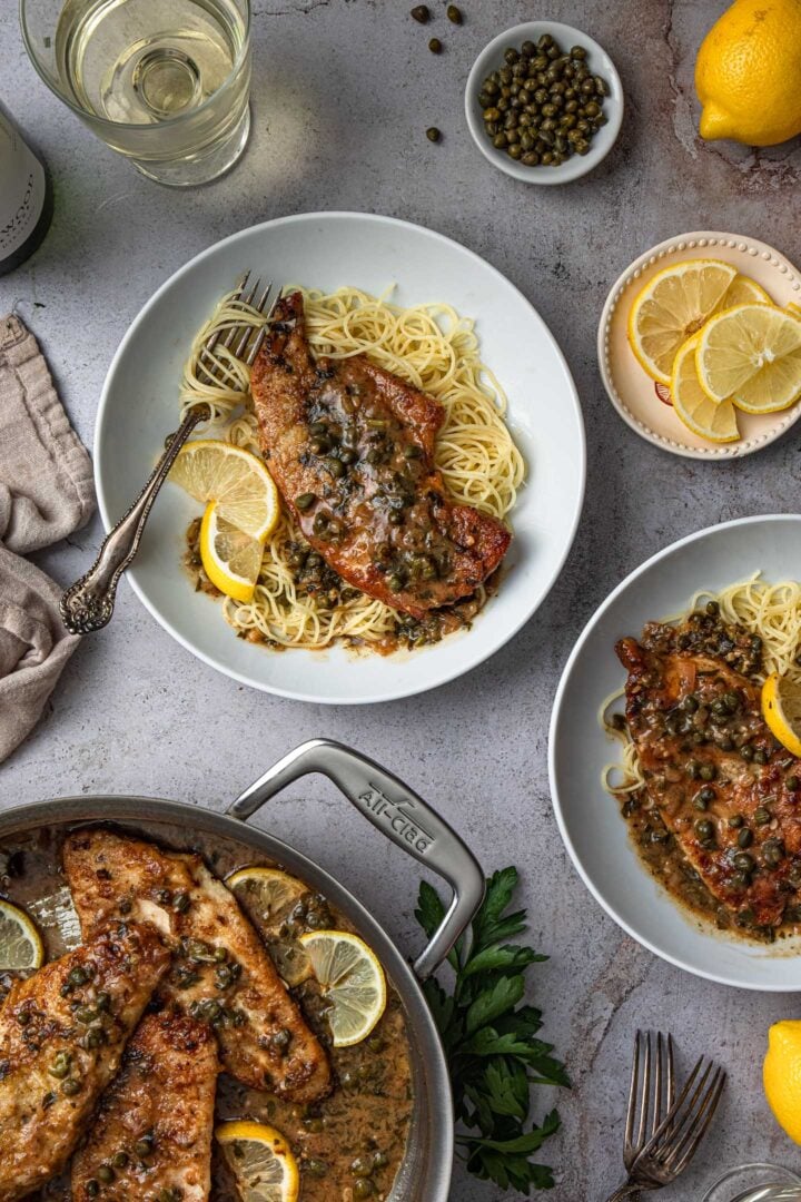 A pan with chicken piccata and two individual servings served over angel hair pasta.