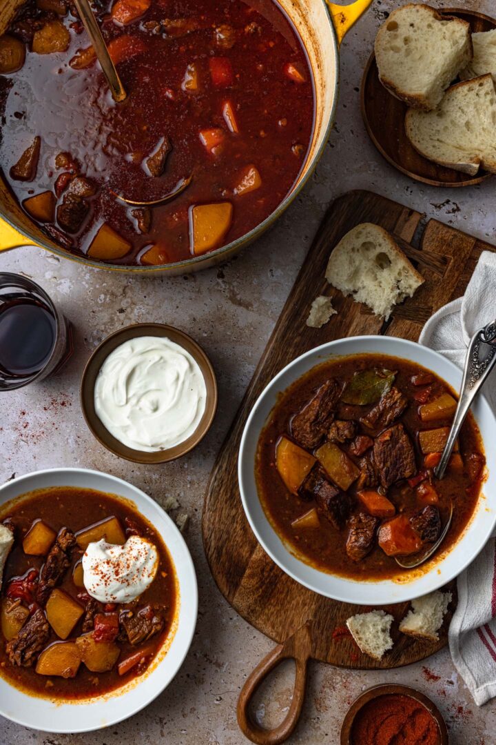 An overhead image of a pot of goulash soup and two bowls with goulash, served with sour cream and bread.