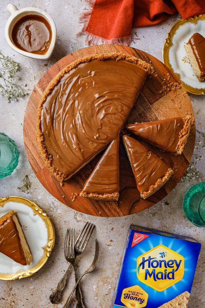 An overhead photo of a dulce de leche cheesecake on a cake stand, two slices of the cheesecake on dessert plates, a bowl of dulce de leche sauce, a box of Honey Maid graham crackers, three forks and two glasses of water.