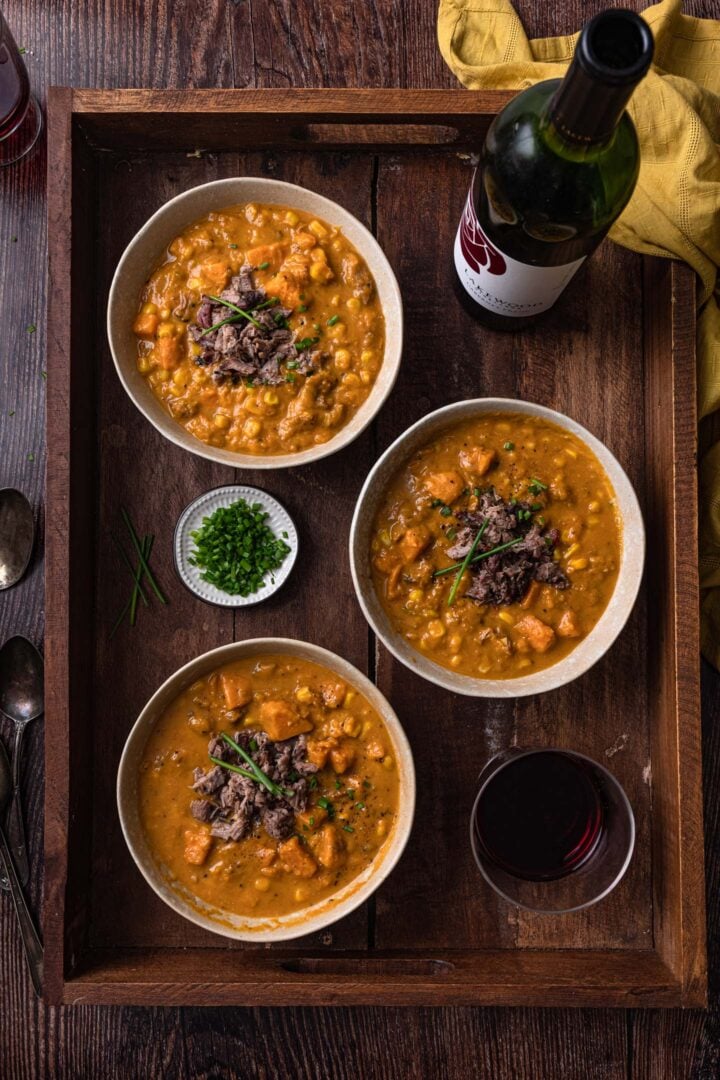 Three bowls of sweet potato chowder on a tray, with a glass of wine, a bottle of wine and a small pinch bowl with chopped chives.