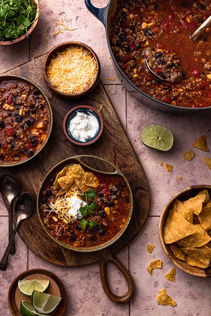 Two bowls of ground beef taco soup on a wood board. Toppings are seen on the side, in bowls, as well as the taco soup pot and two spoons.