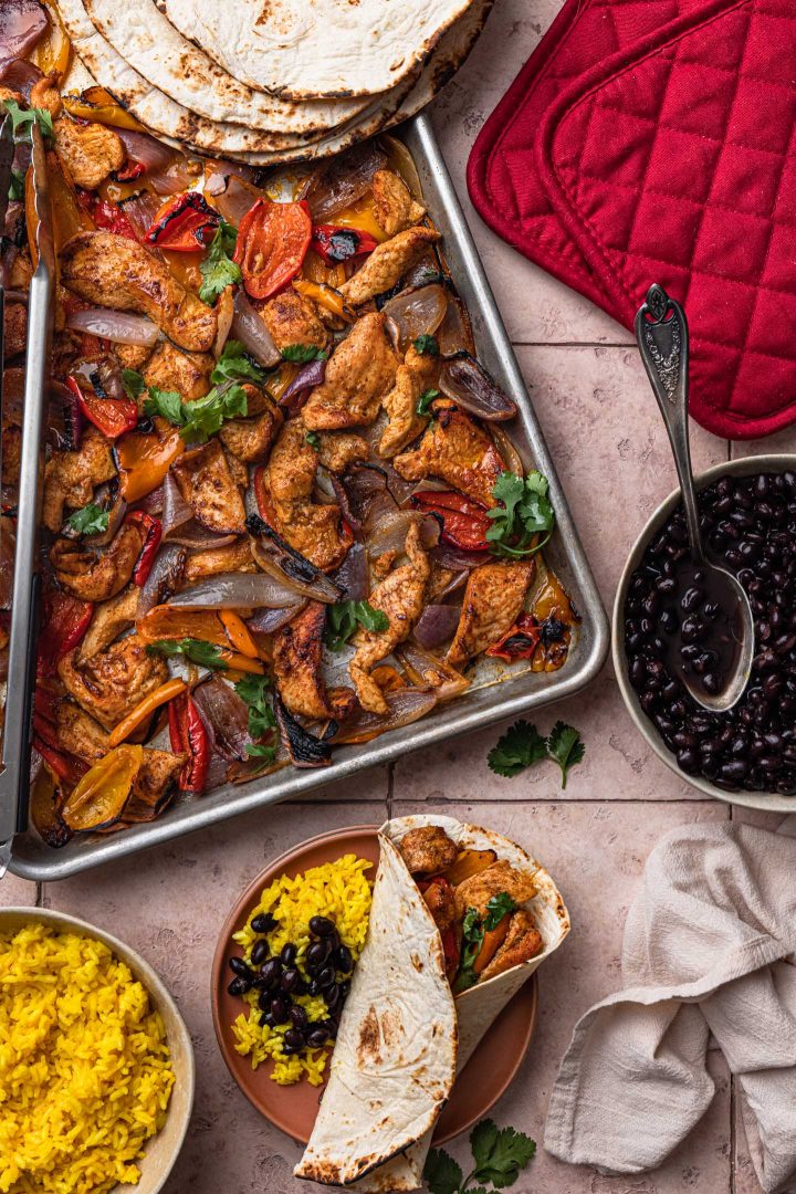 A serving scene: the fajitas in a baking sheet, a bowl of black beans, a bowl of rice and an assembled fajita in a tortilla.