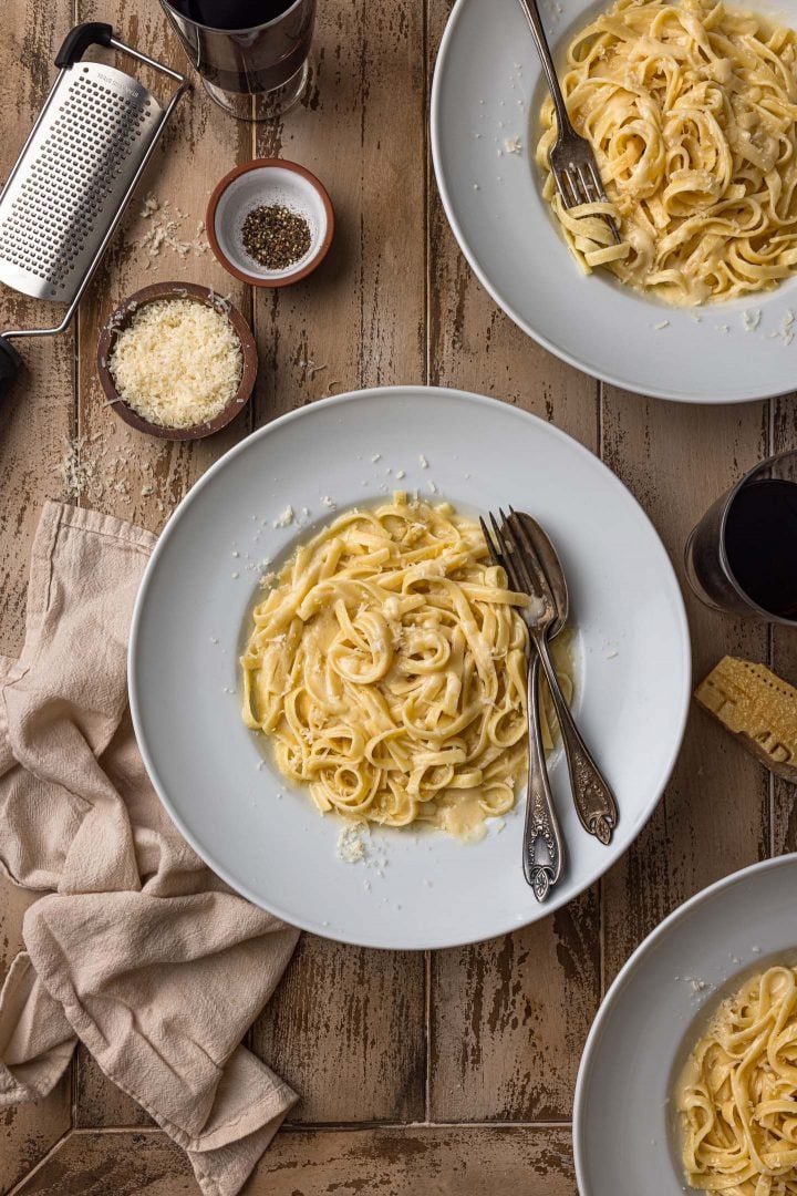 An overhead photo of three bowls of the original fettuccine Alfredo, a smaller bowl with parmesan and one with black pepper, a cheese grater, a parmesan rind and two glasses of red wine.