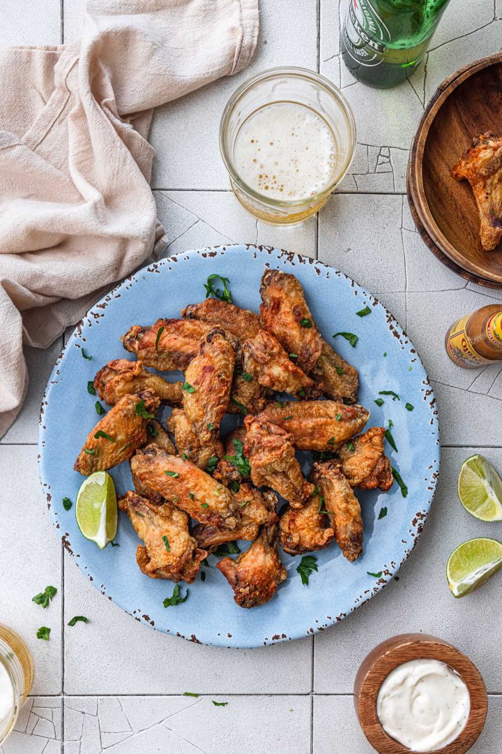 An overhead photo of a platter of air fryer chicken wings, served with lime wedges, a dipping sauce, a hot sauce bottle and beer.