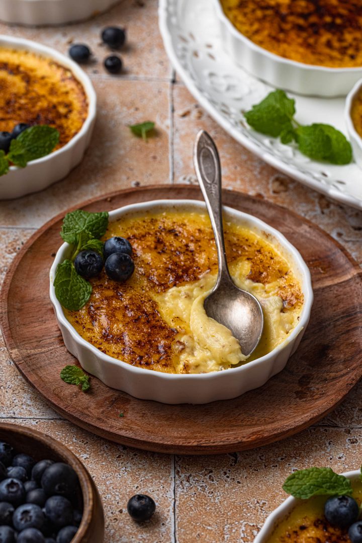 Vanilla creme brulee in a ramekin, with a spoon showing the creamy custard. The creme brulee is garnished with mint and blueberries.