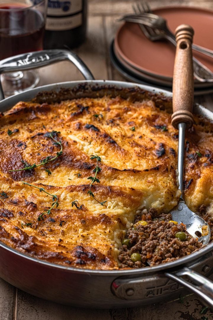 Shepherd's Pie in a pan, garnished with thyme. A spoon is scooping some of the lamb filling.