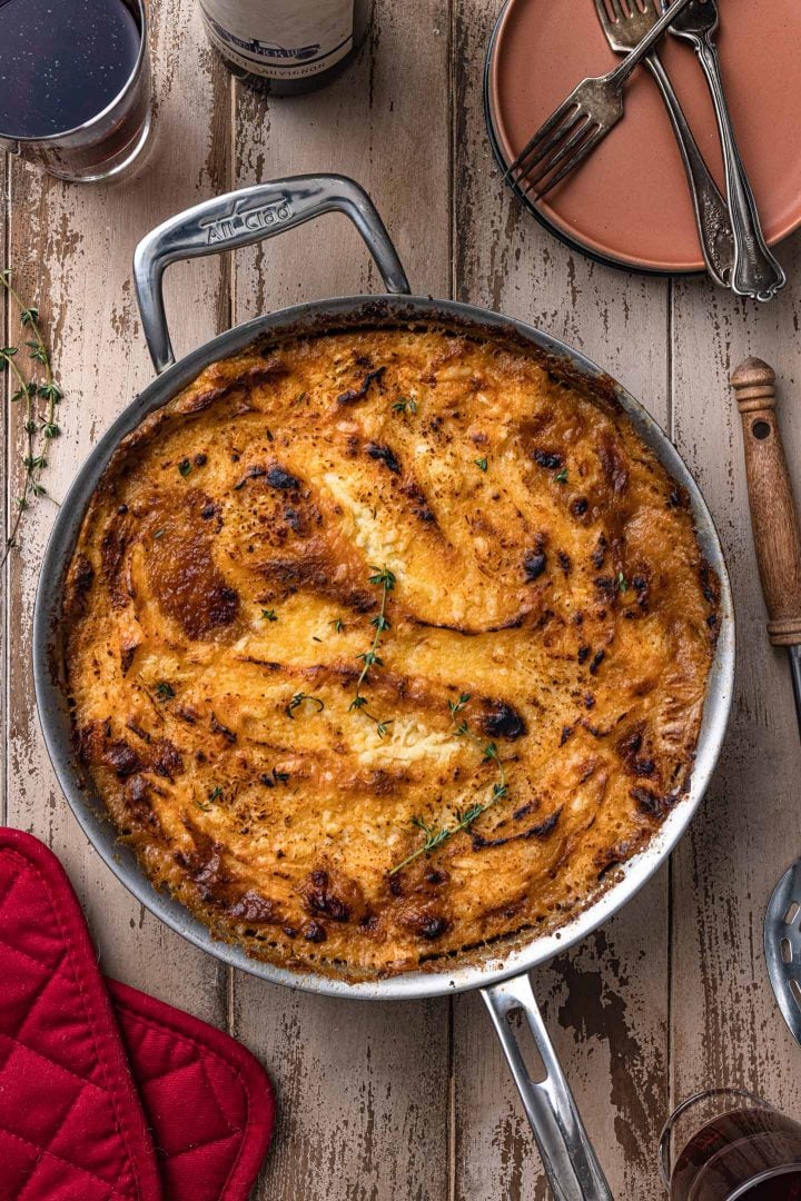 An overhead photo of a shepherd's pie, with a serving spoon, some pla
tes and forks, and two oven mitts.