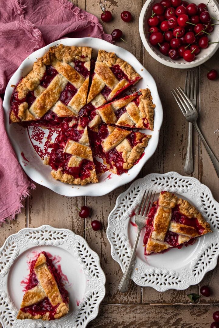 A serving scene: A few slices in plates and some slices still in the pie dish. You can also see a bowl of fresh sour cherries, a few forks and a napkin.