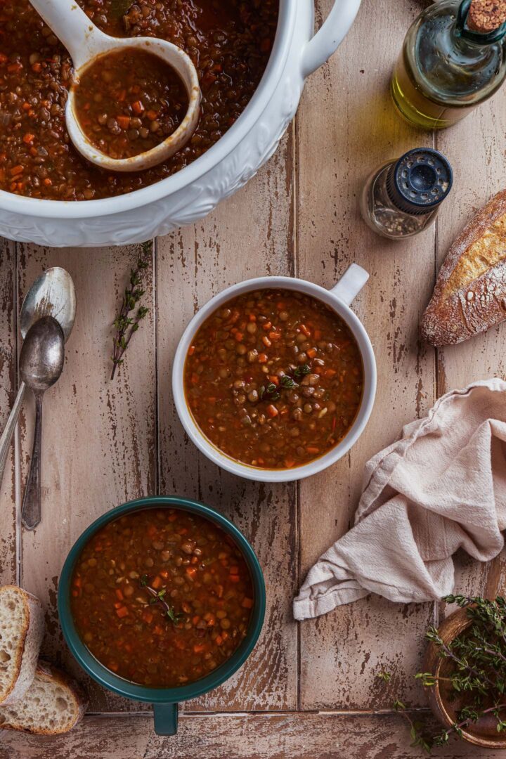A serving scene: you can see the soup tureen, as well as two bowls of lentil soup. There are also spoons, fresh thyme, a kitchen towel, crusty bread, pepper and an olive oil jar.