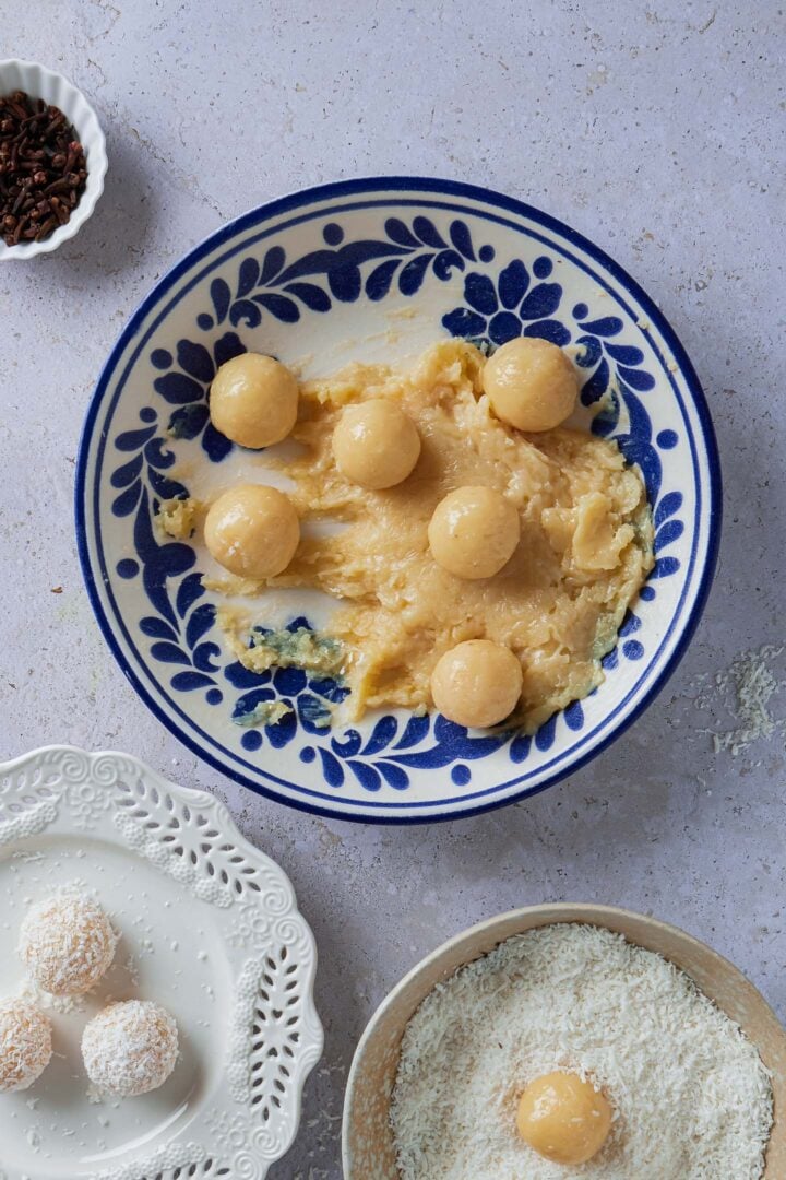A plate with the beijinho mixture, with some beijinho balls. You can also see a small bowl with whole cloves, a plate with grated coconut and a plate with a few fully assembled beijinhos.