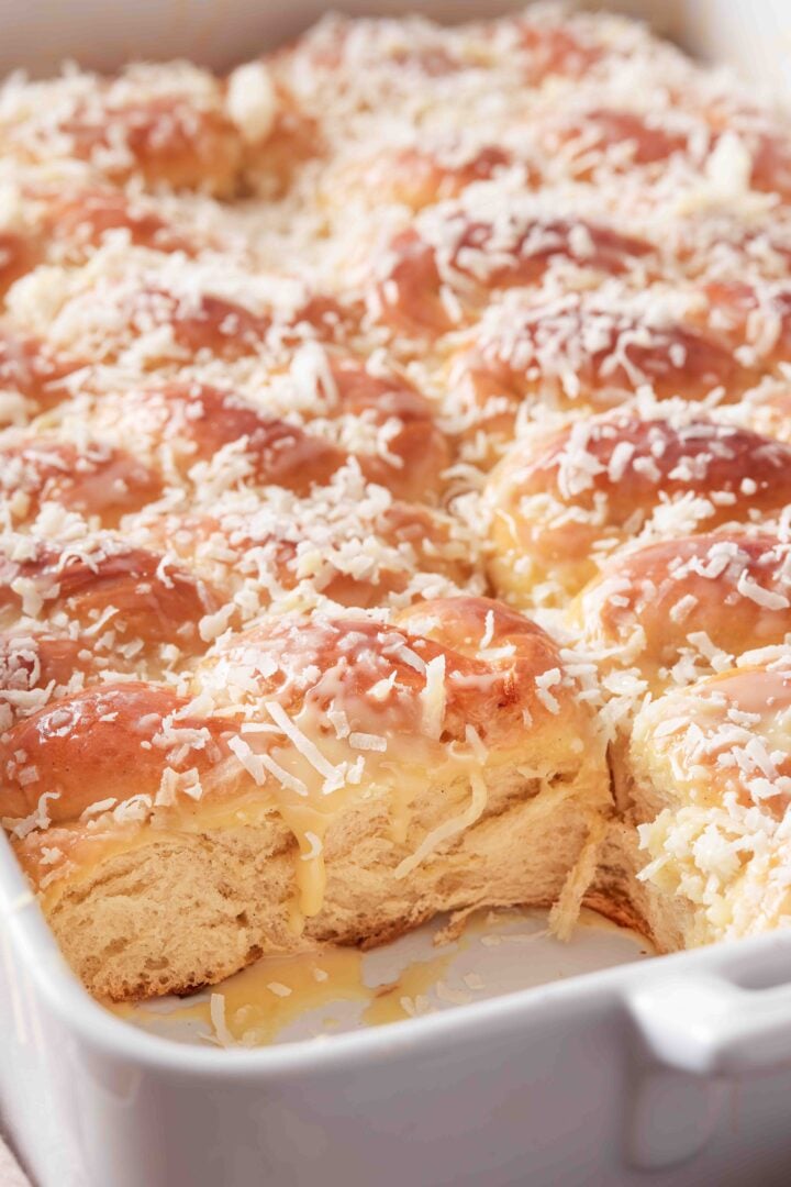 Close-up of Brazilian sweet condensed milk buns in a white baking dish, topped with shredded coconut and drizzled with sweetened condensed milk.