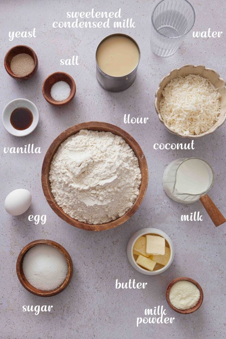Overhead photo of the ingredients for Brazilian sweet condensed milk buns, including flour, milk, sweetened condensed milk, coconut, sugar, butter, egg, vanilla, yeast, salt, water, and milk powder.