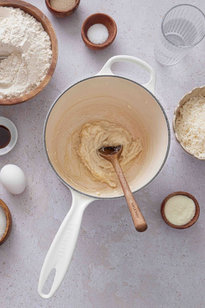 Overhead photo of the cooked tangzhong paste in a white saucepan with a wooden spoon, surrounded by ingredients for Brazilian sweet condensed milk buns.