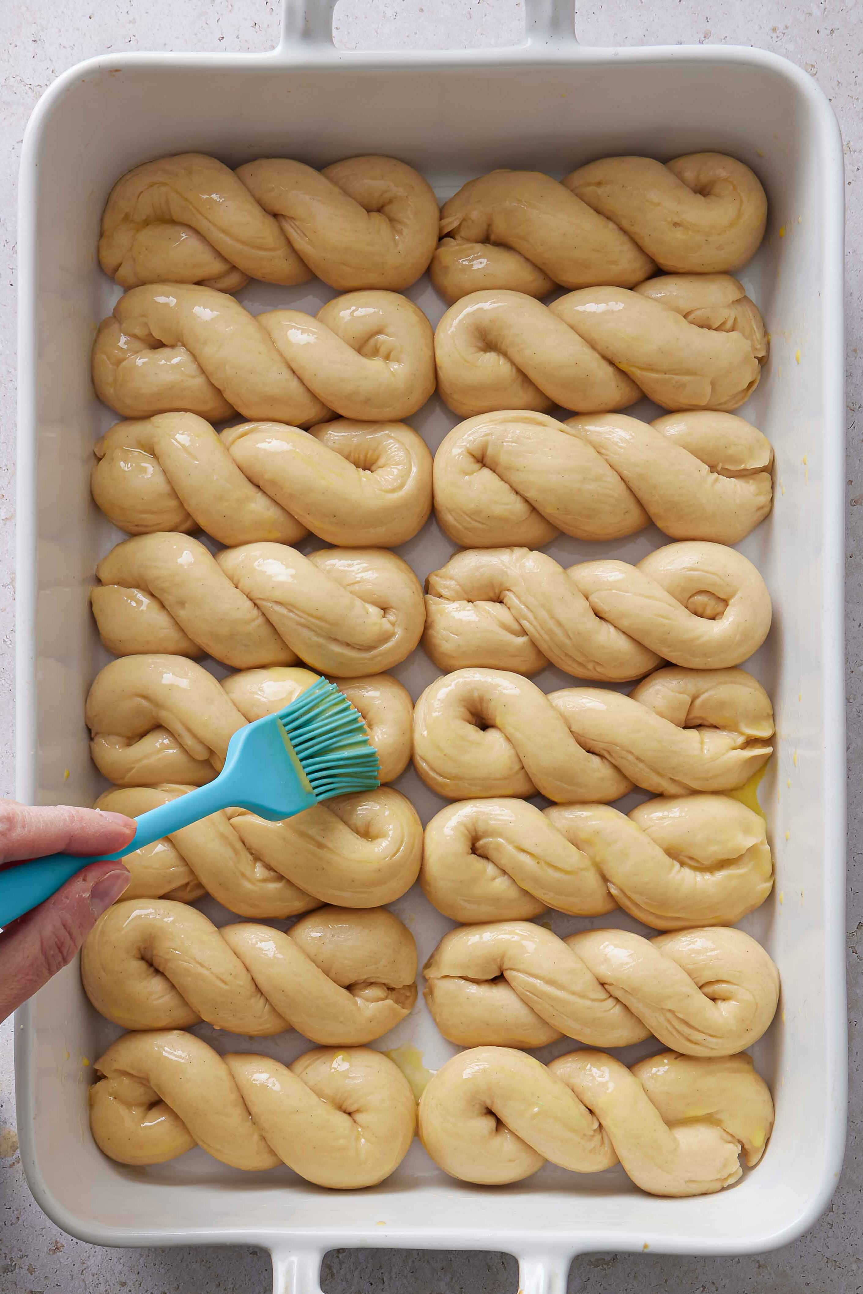 Overhead photo of egg wash being brushed over the risen buns in the baking dish before baking.