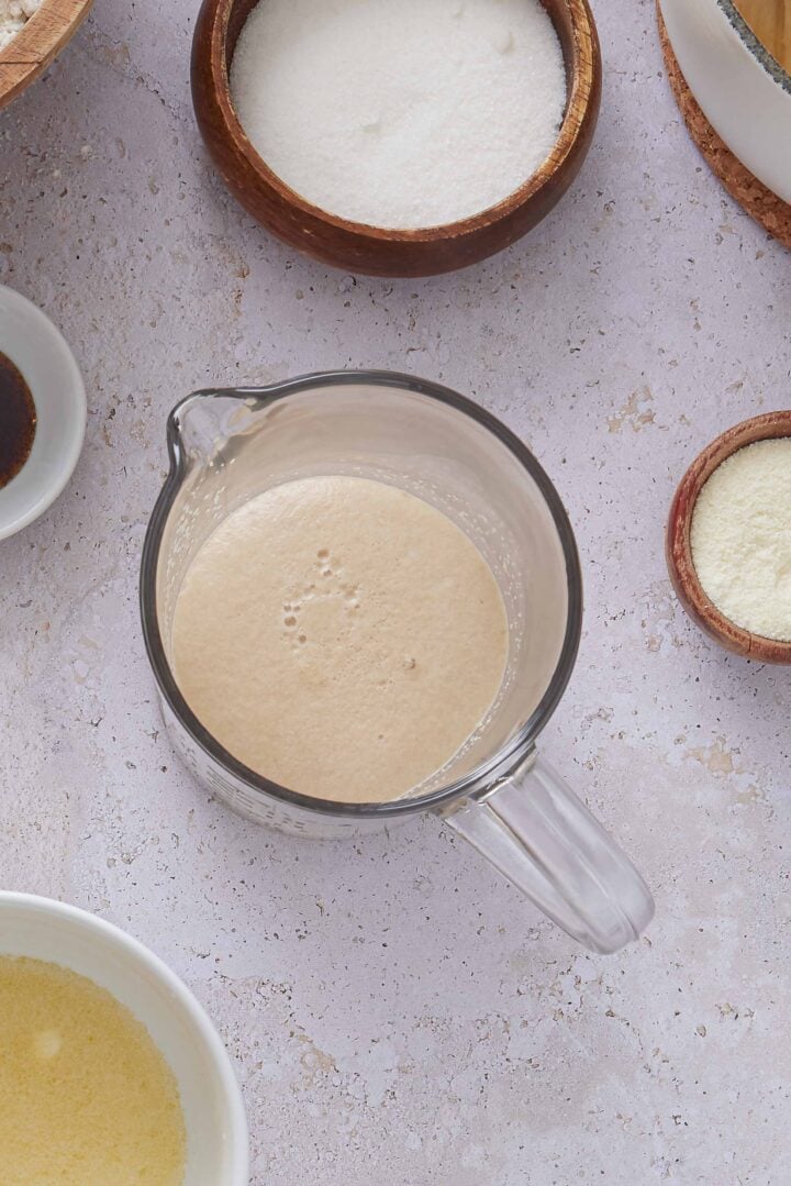 Overhead photo of foamy yeast mixture in a glass measuring cup, surrounded by ingredients for Brazilian sweet condensed milk buns.